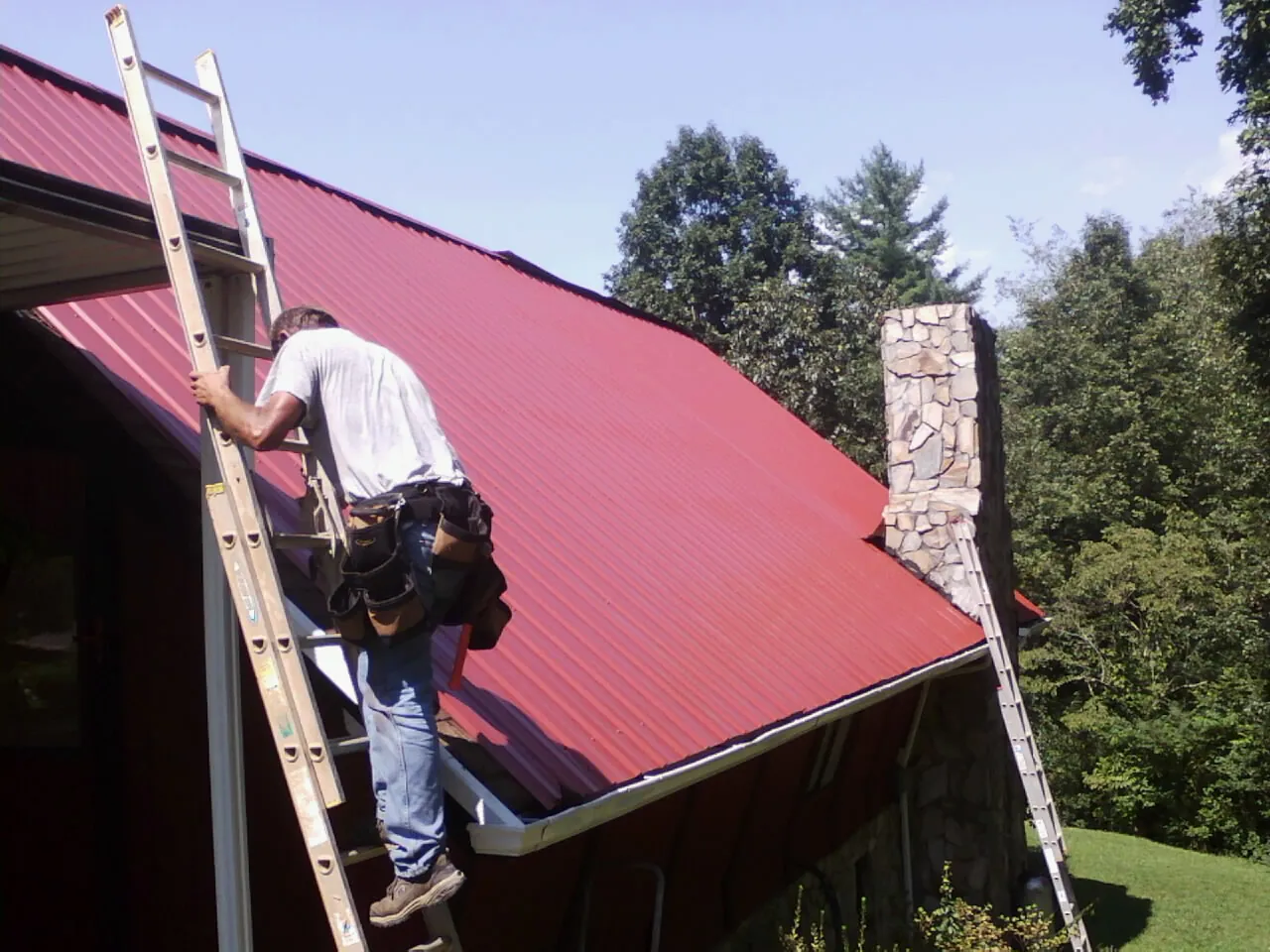 Roofer on a ladder beside a red metal roof and stone chimney — Soffit & Fascia Repair by Roofing Brothers in Meacham